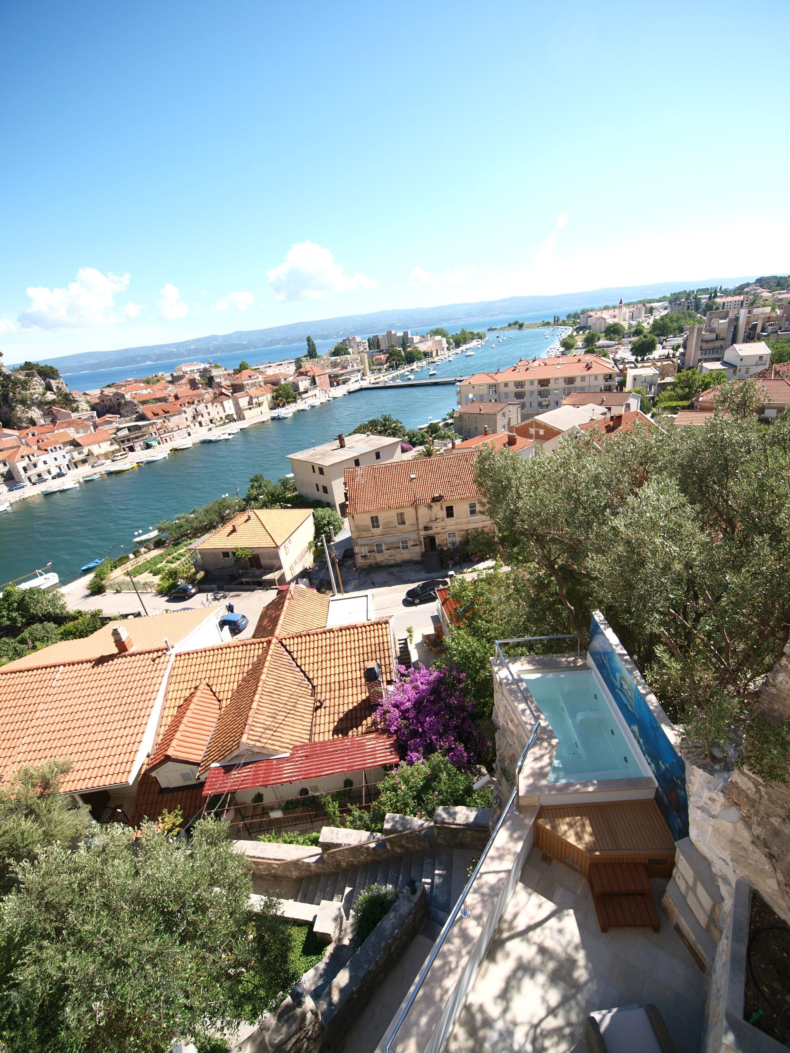 A coastal town with terracotta-roofed buildings, a marina, and a swimming pool.