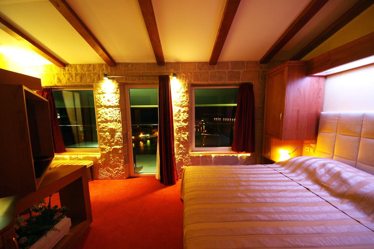 Habitación de hotel con cama, televisor, una mesita con una planta y vistas a la ciudad desde las ventanas.