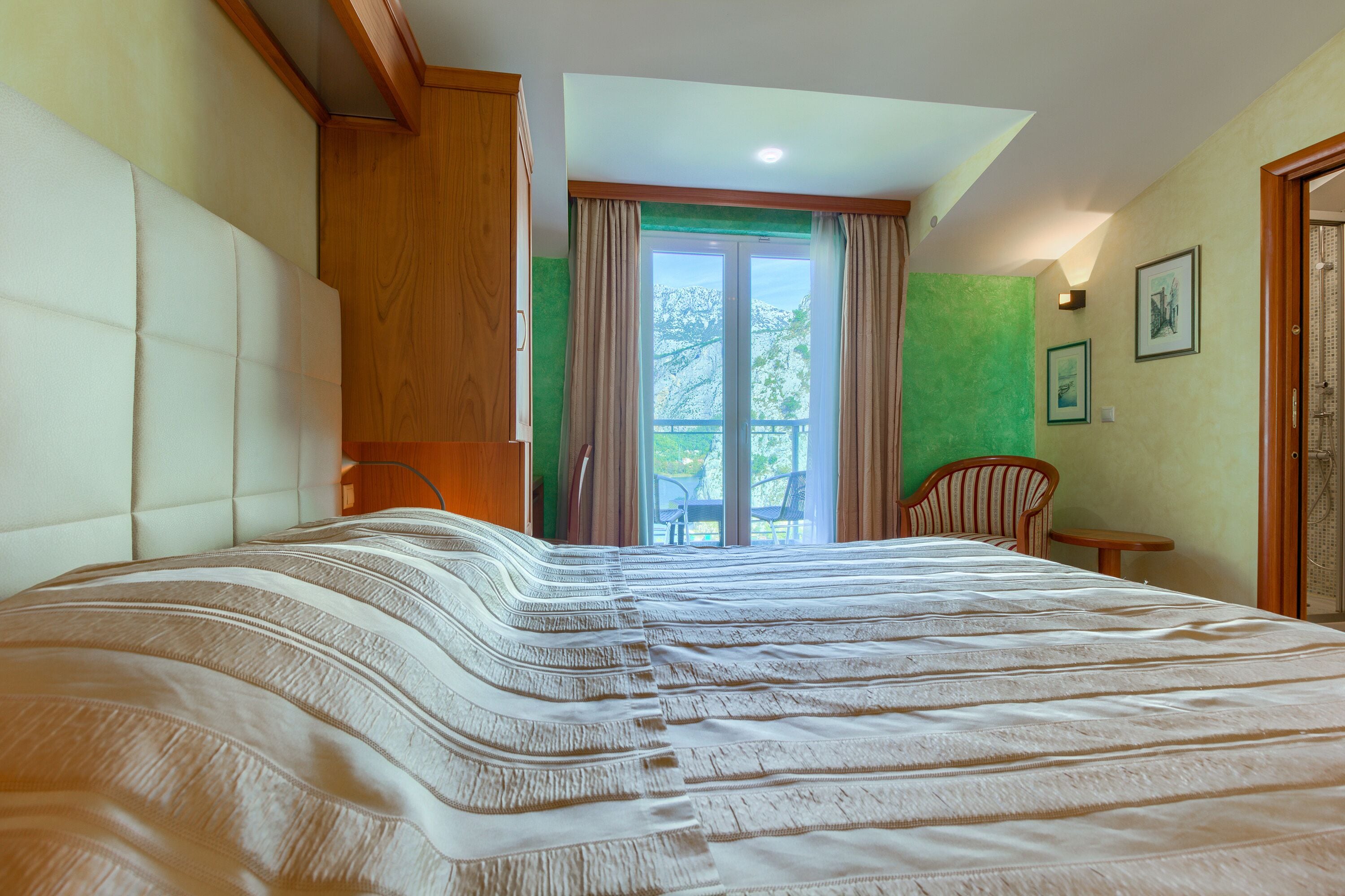 A bedroom with a large bed, a wooden wardrobe, a chair, a small table, and a balcony with a view.