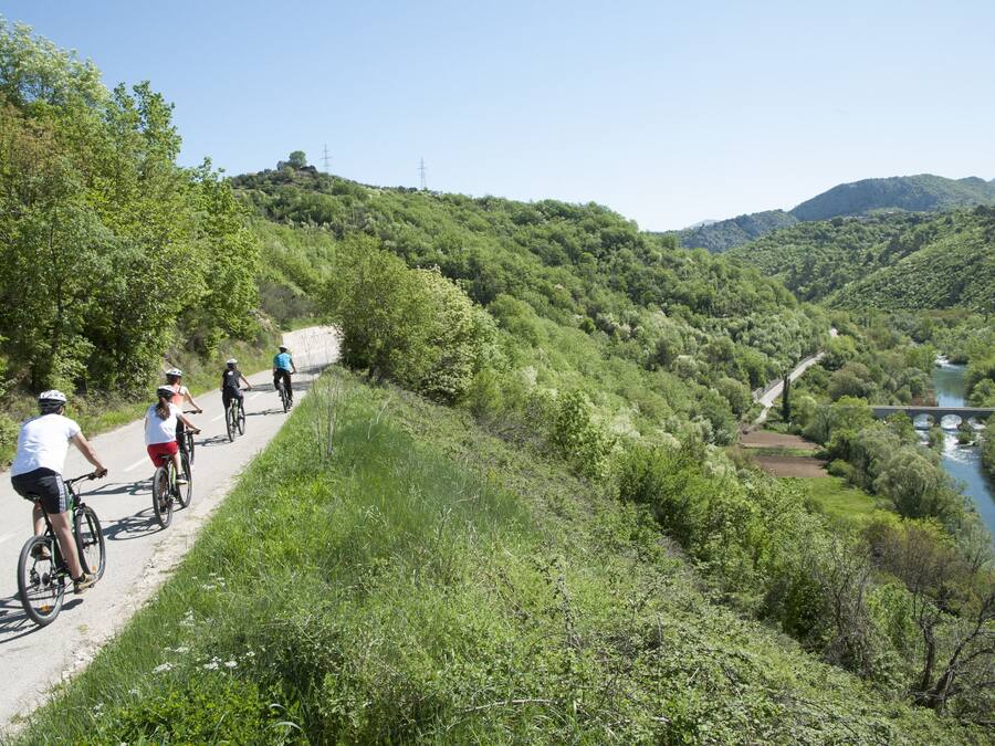 Un grupo de ciclistas en un camino pavimentado junto a un río, con un puente al fondo.