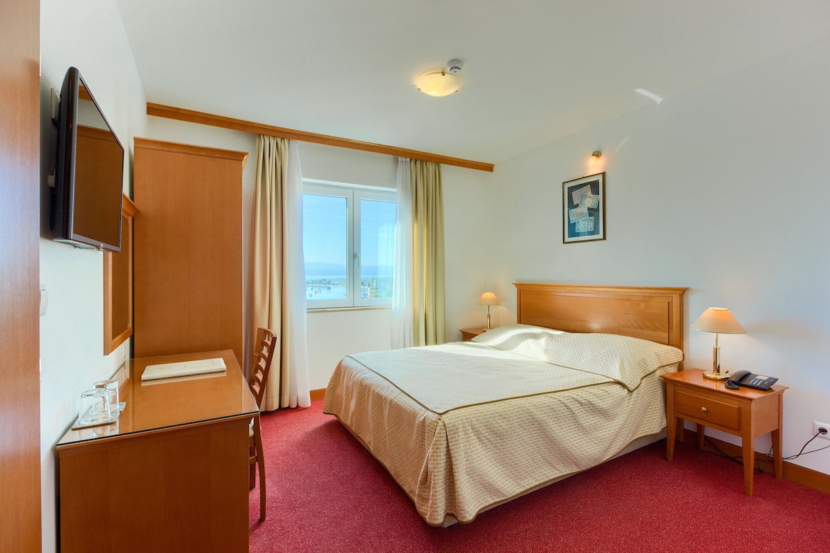 Habitación de hotel con cama, mesita de noche con lámparas, escritorio y silla, televisor y ventana con cortinas.