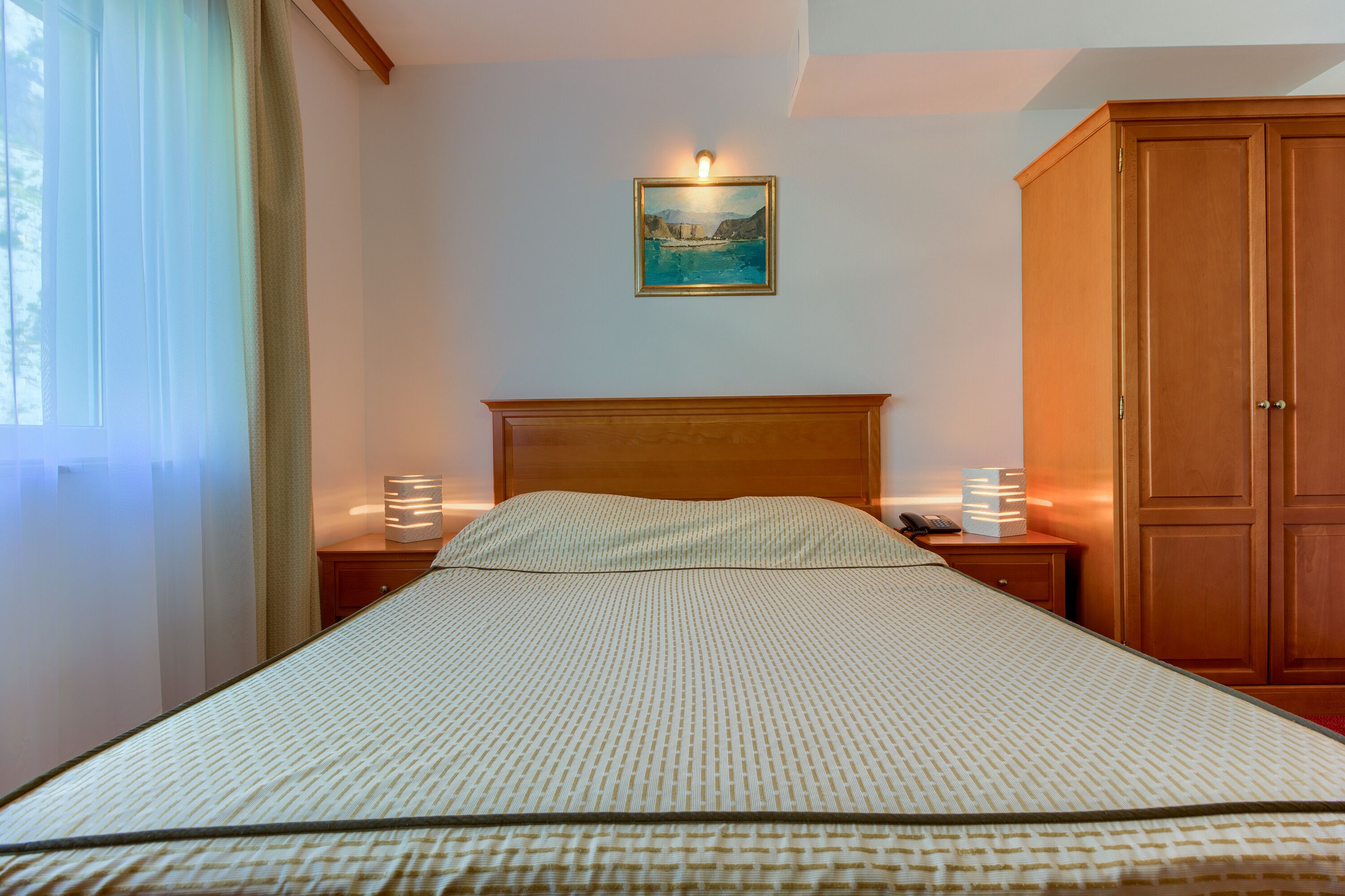 A neatly made bed with a patterned bedspread, a wooden headboard, bedside lamps, a nightstand, and a wooden wardrobe.