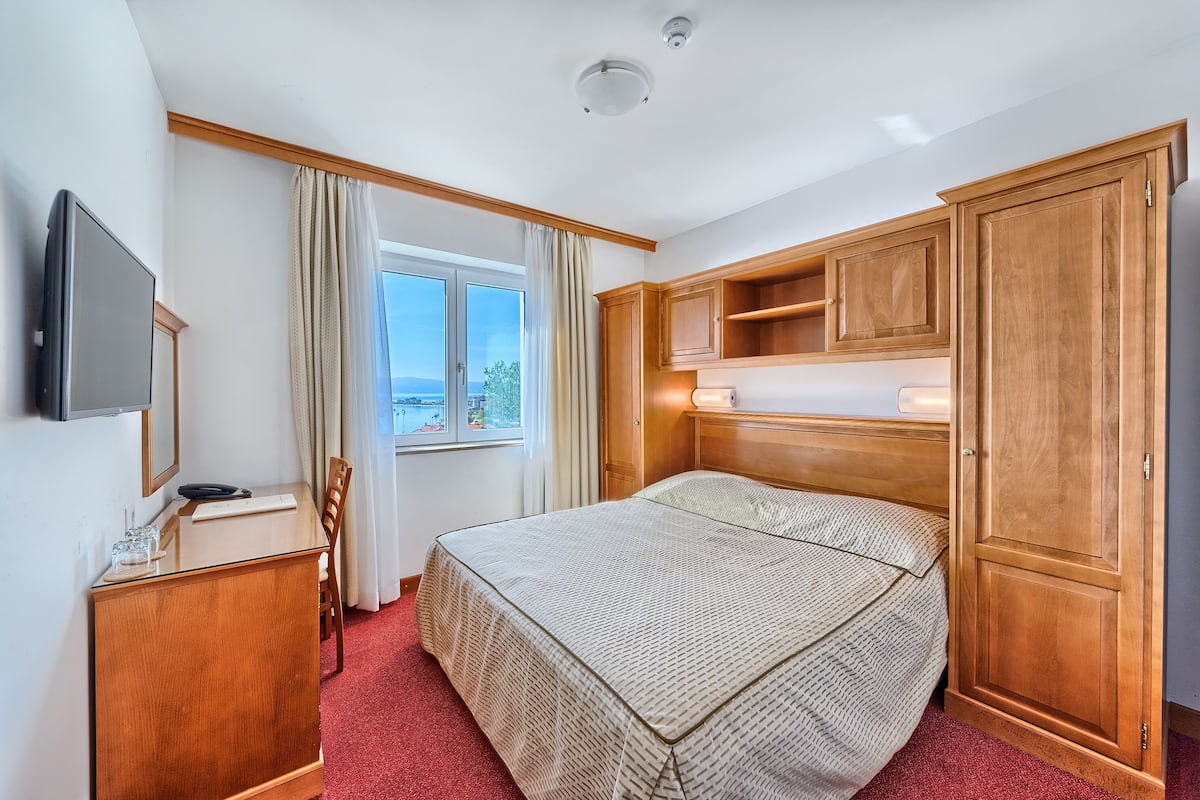 Habitación de hotel con cama, armario de madera, escritorio con silla, televisor y ventana con cortinas.