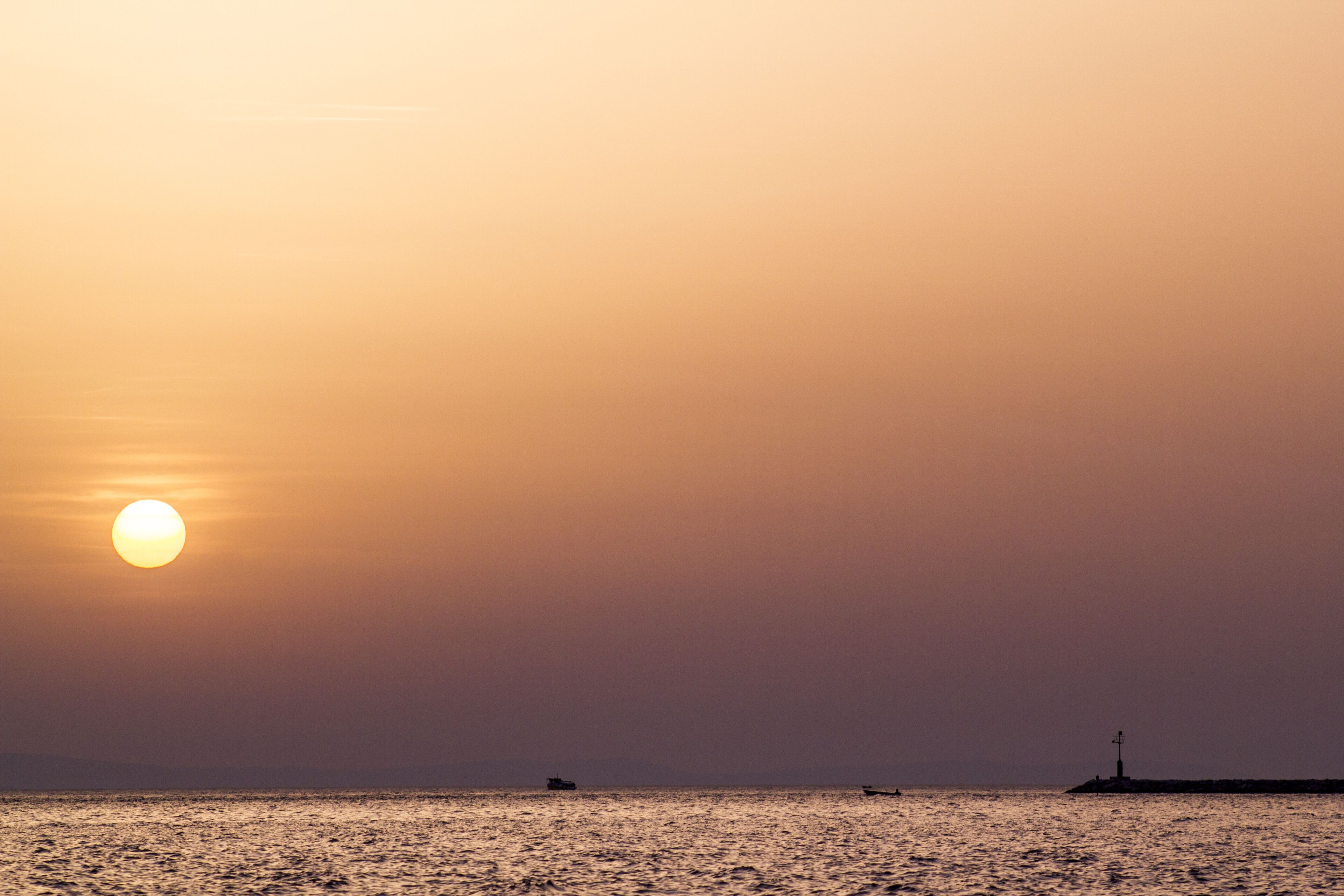 A sunset over a calm sea with a distant lighthouse and a small boat.