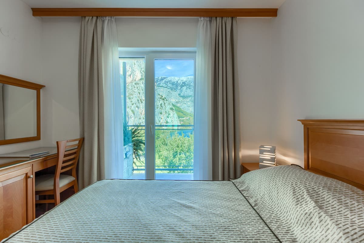 Habitación de hotel con cama, escritorio, silla, mesita de noche y vista a un paisaje montañoso a través de un ventanal grande.