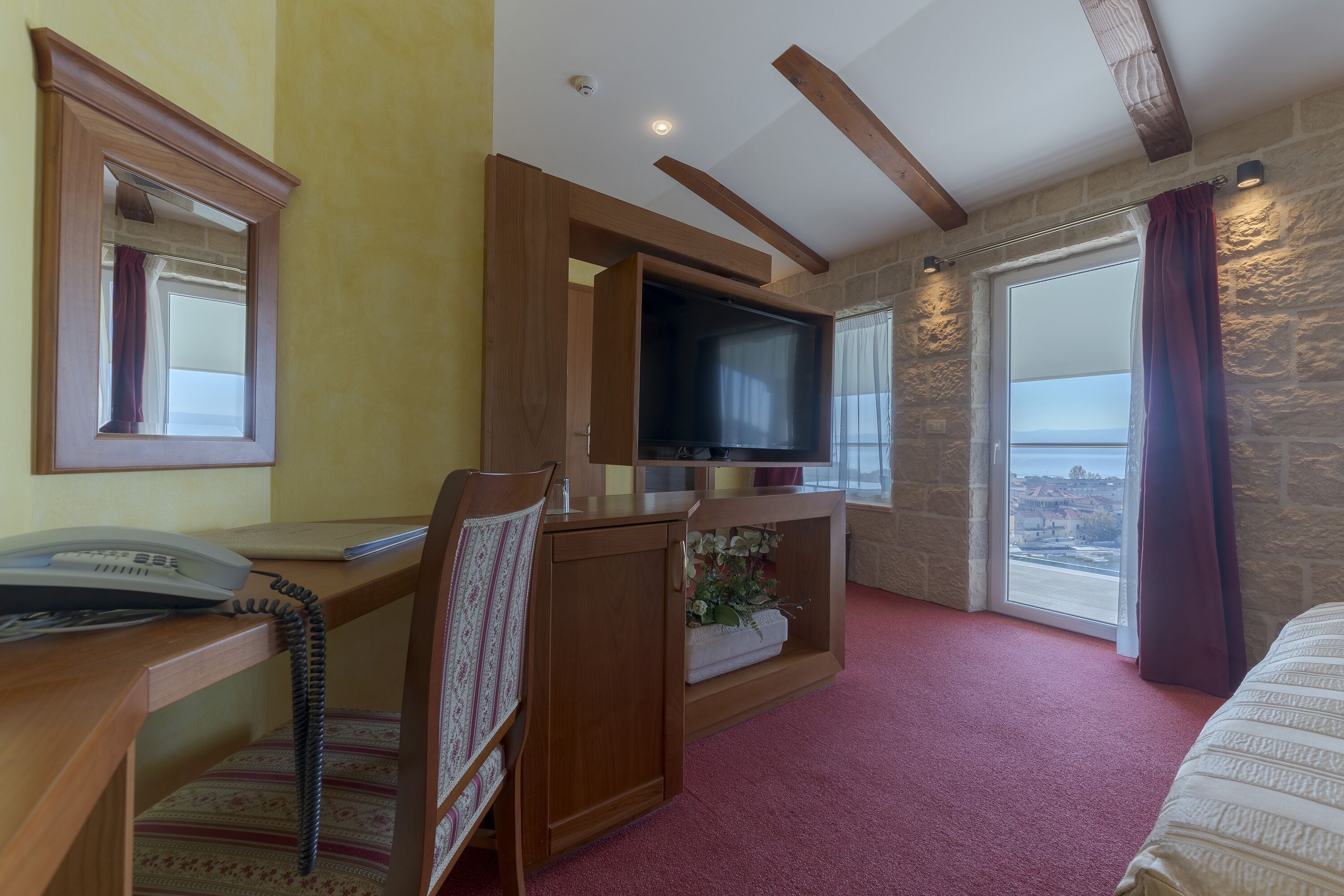 A hotel room with a wooden desk, a telephone, a television, a cushioned chair, a small plant, and a large window with a view.