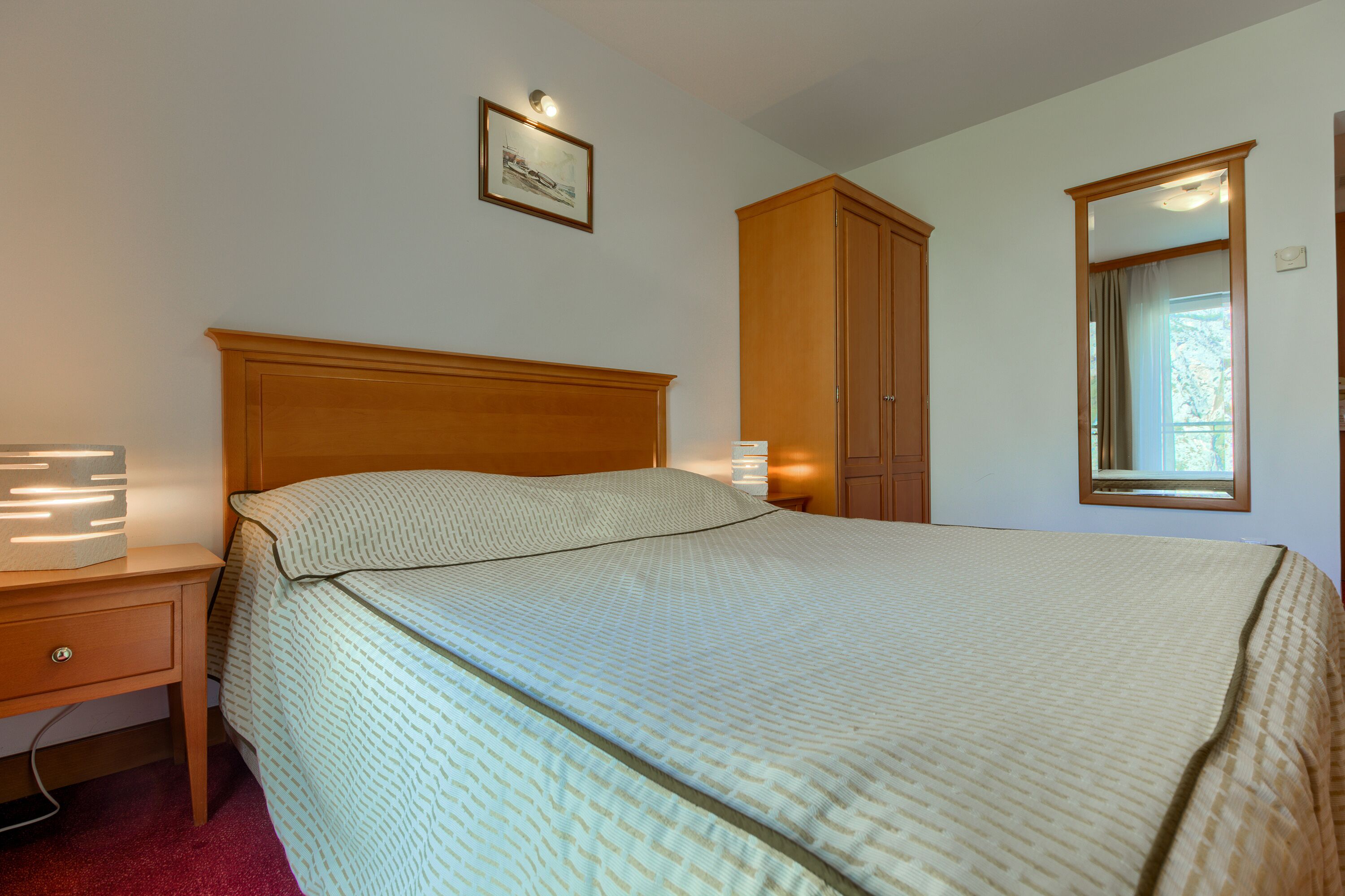 A bedroom with a bed, a nightstand, a wooden wardrobe, a framed picture on the wall, and a window with curtains.