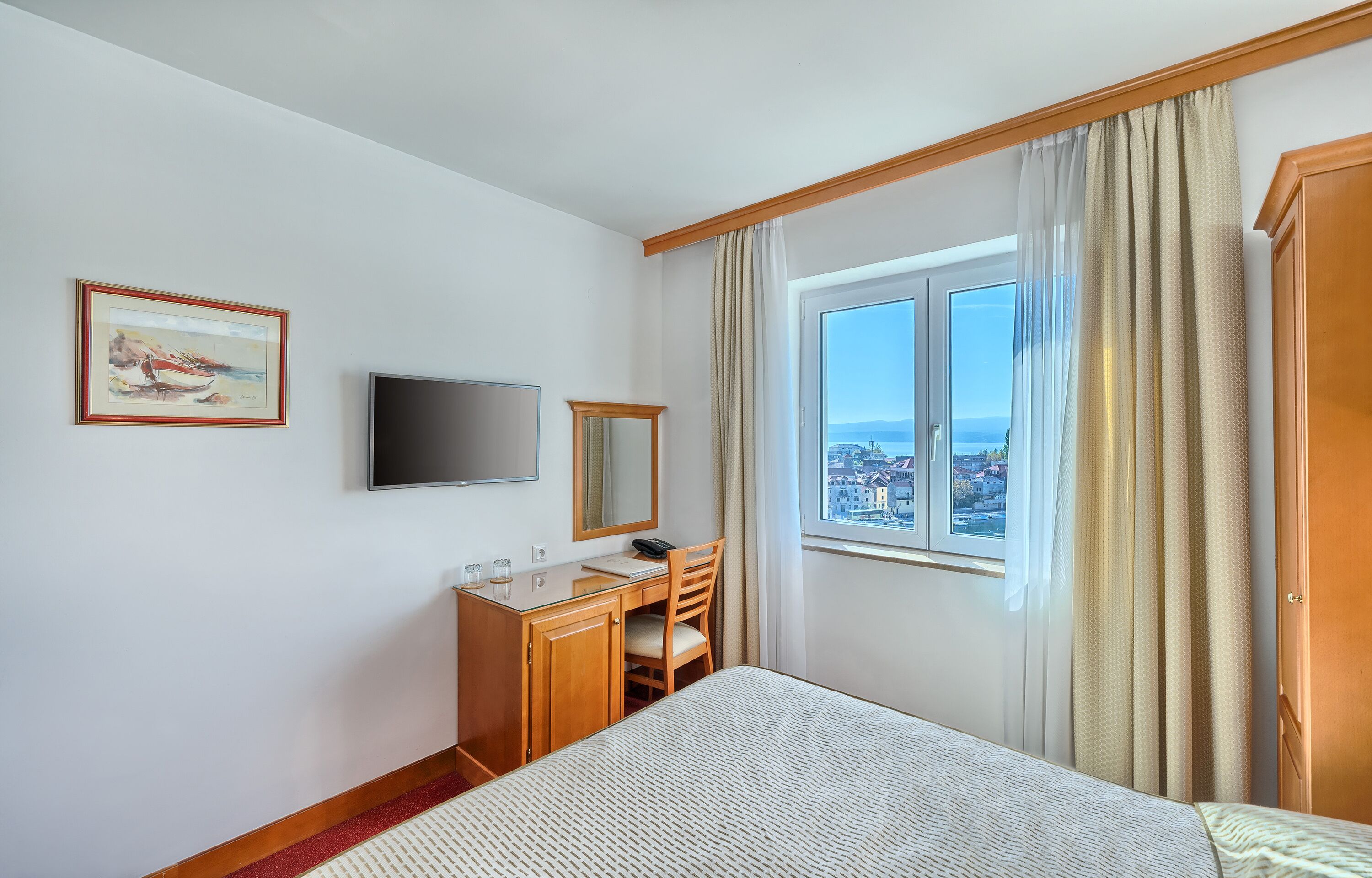 Habitación de hotel con cama, escritorio con silla, televisor, ventana con cortinas y un cuadro en la pared.