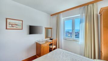 A hotel room with a bed, a desk with a chair, a television, a window with curtains, and a painting on the wall.