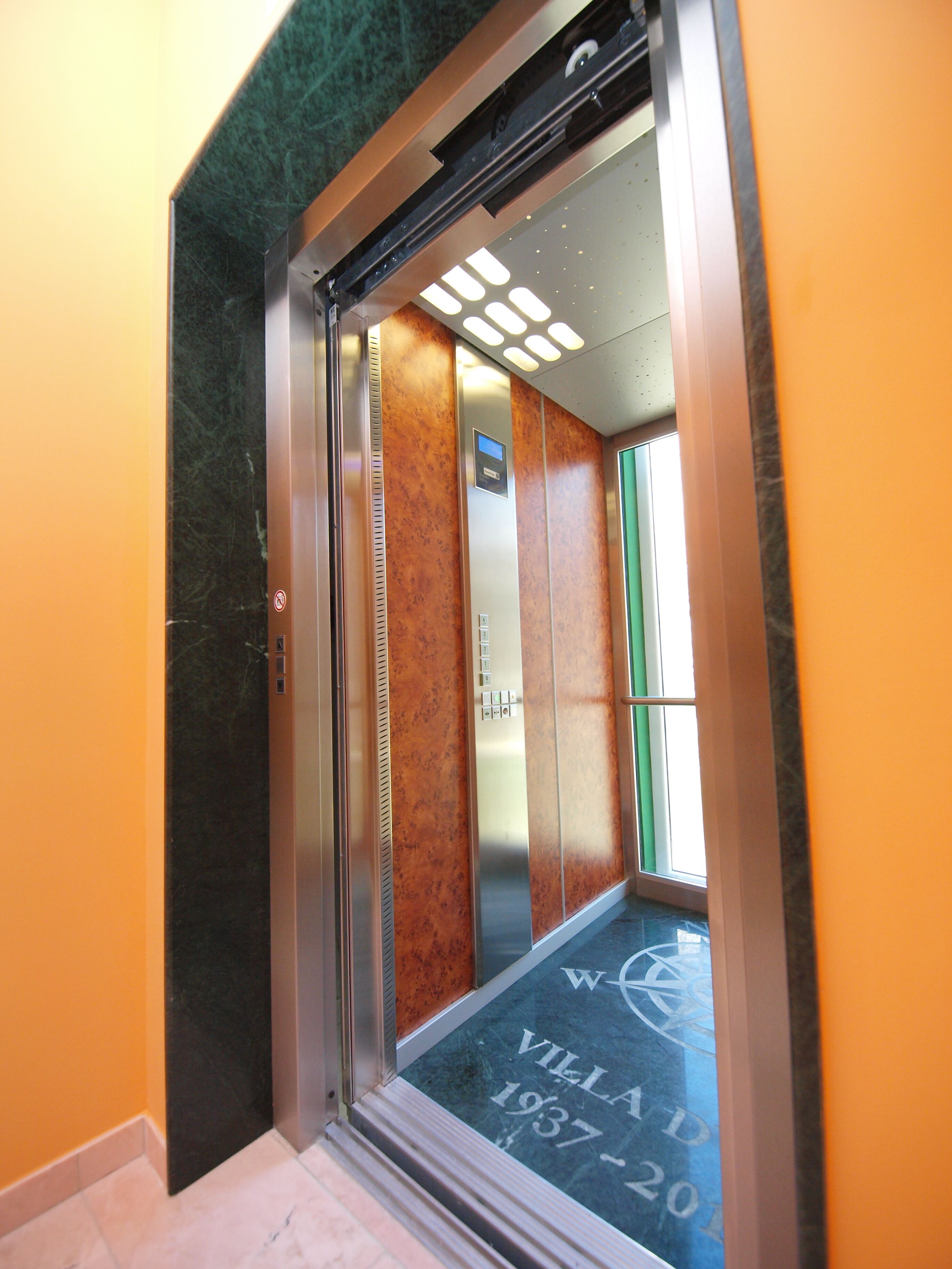 Interior d'un ascensor amb paret de marbre, porta verda i sòl gravat amb la inscripció 'Villa 1937-20'.