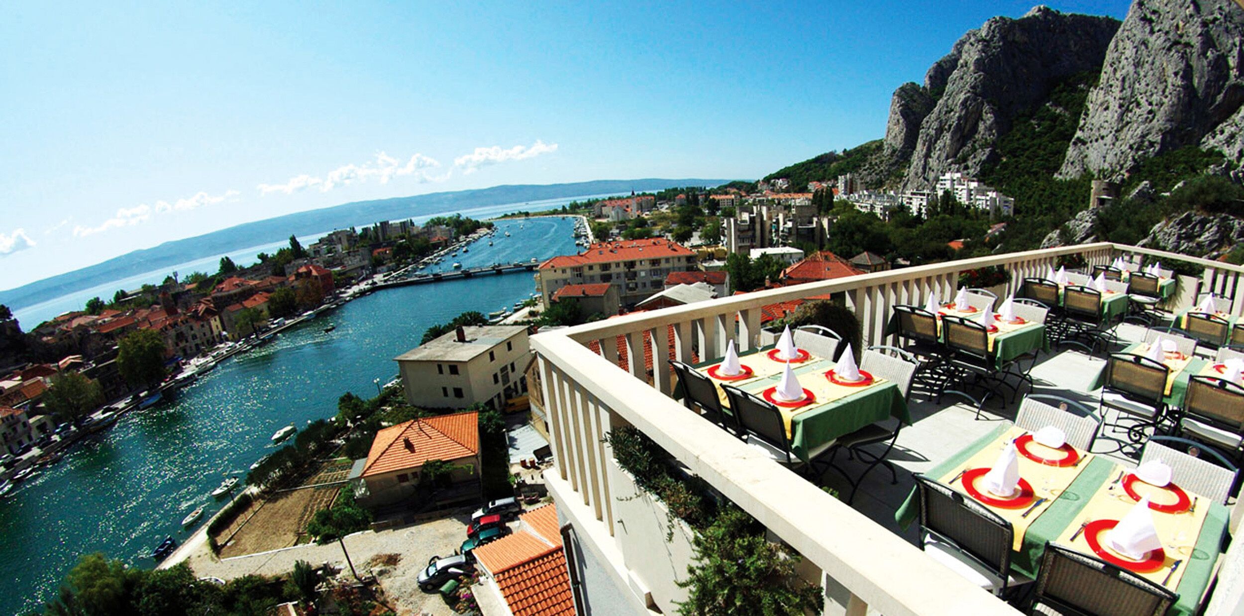 Un hôtel situé sur la côte, doté d’une terrasse où l’on peut prendre ses repas, offrant une vue sur la mer et entouré de bâtiments.