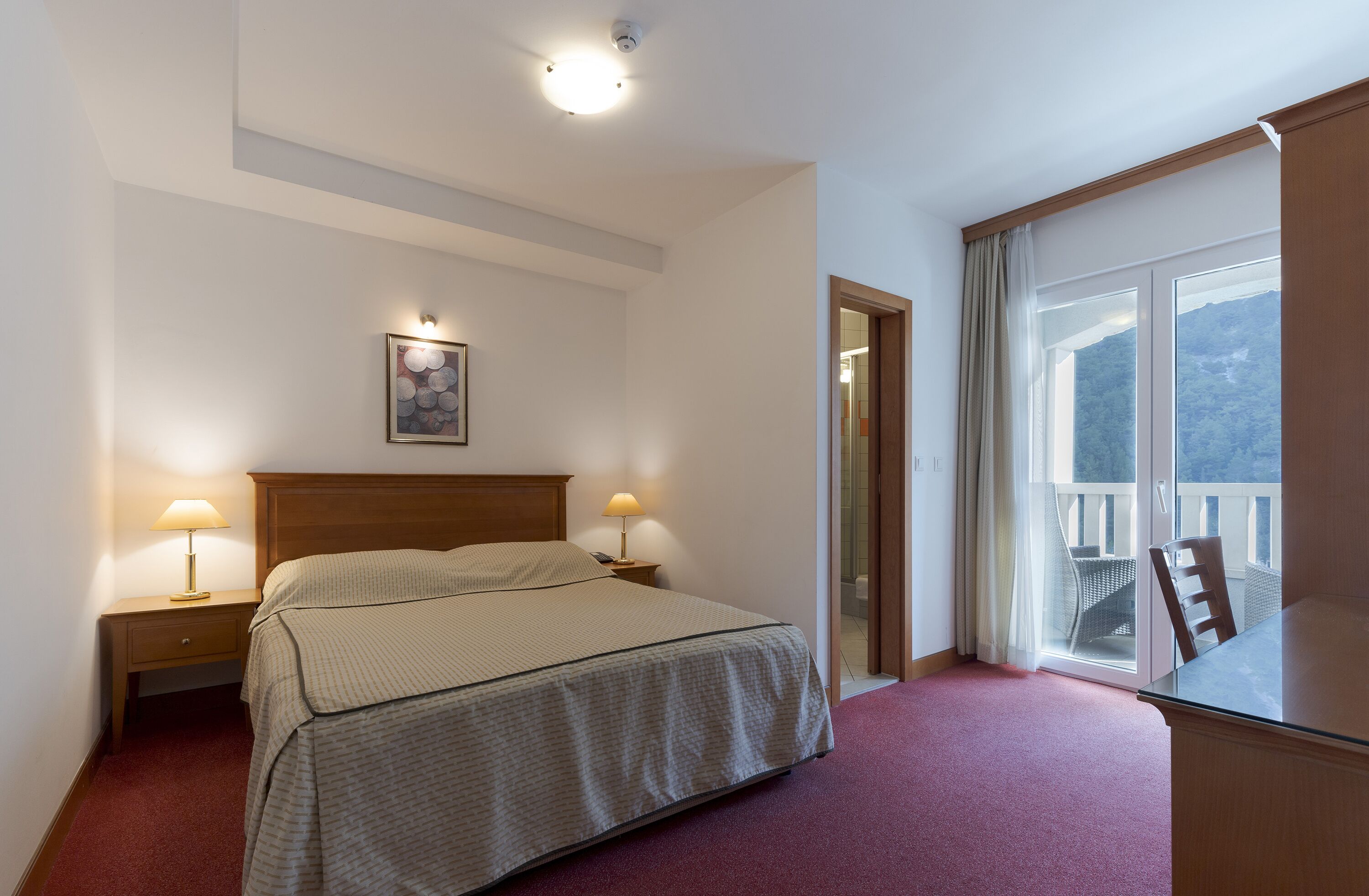 A hotel room with a bed, two bedside lamps, a painting on the wall, a window with a view, and a balcony with chairs.