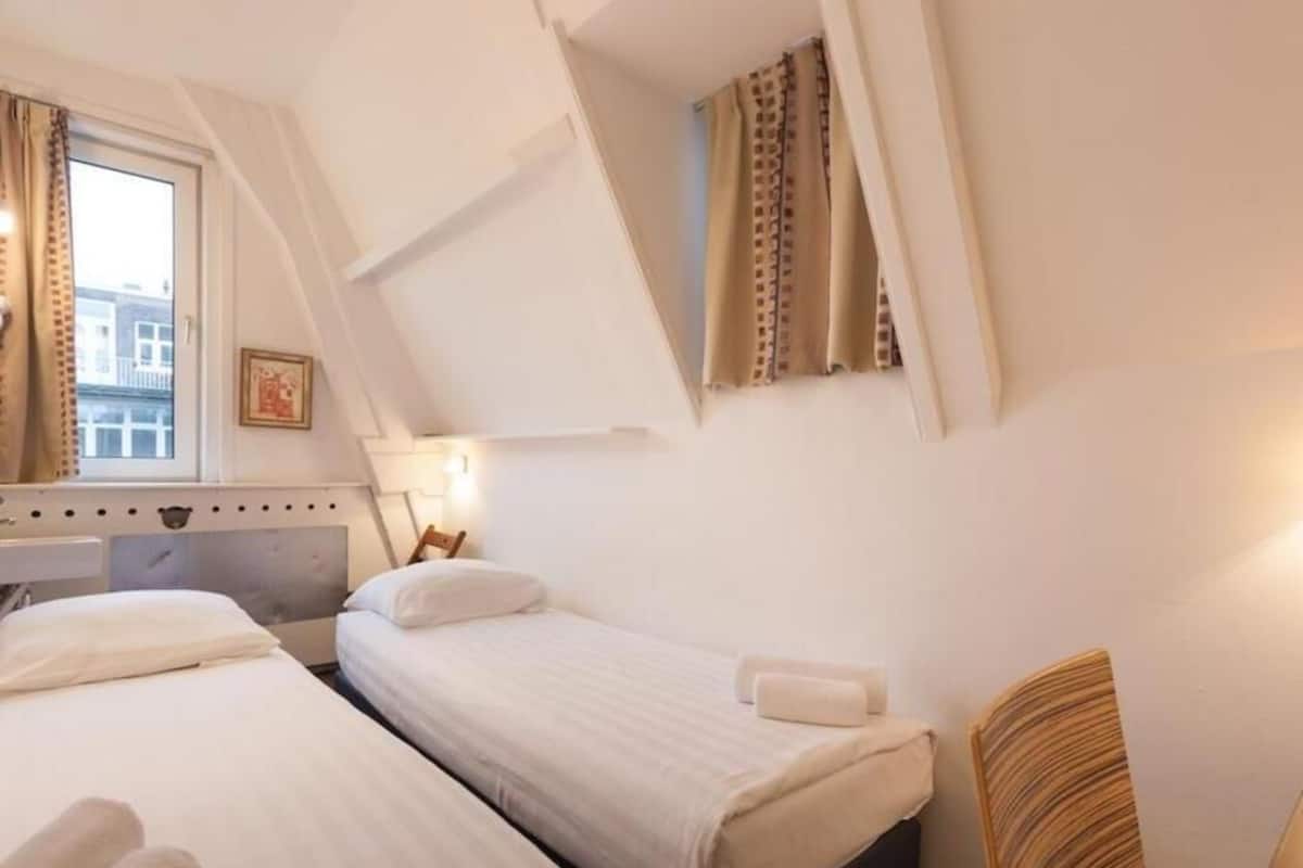 A hotel room with two beds, a window with curtains, a bedside lamp, and a chair.