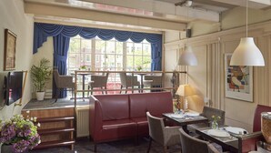 A hotel lobby with a red leather sofa, dining area with tables and chairs, a large window with blue curtains, and a television mounted on the wall. - ITC Hotel (Amsterdam)