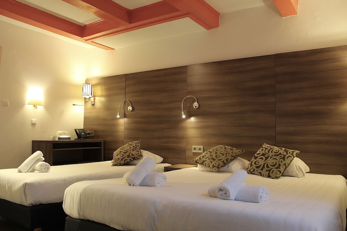 A hotel room with two beds, each with a rolled towel, wooden headboard, and patterned pillows. There are wall-mounted lamps and a small table with a phone and a lamp.