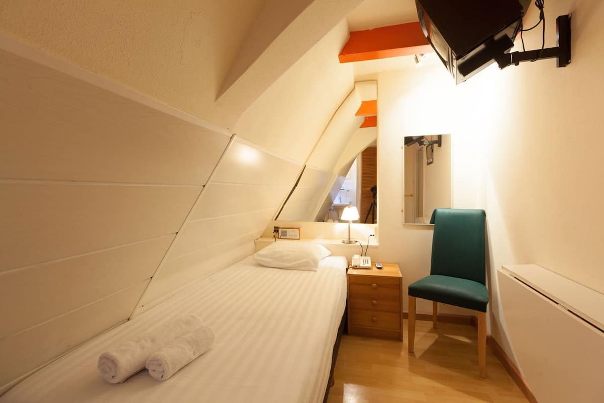 A small, triangular room with a bed, a chair, a nightstand, and a wall-mounted TV.