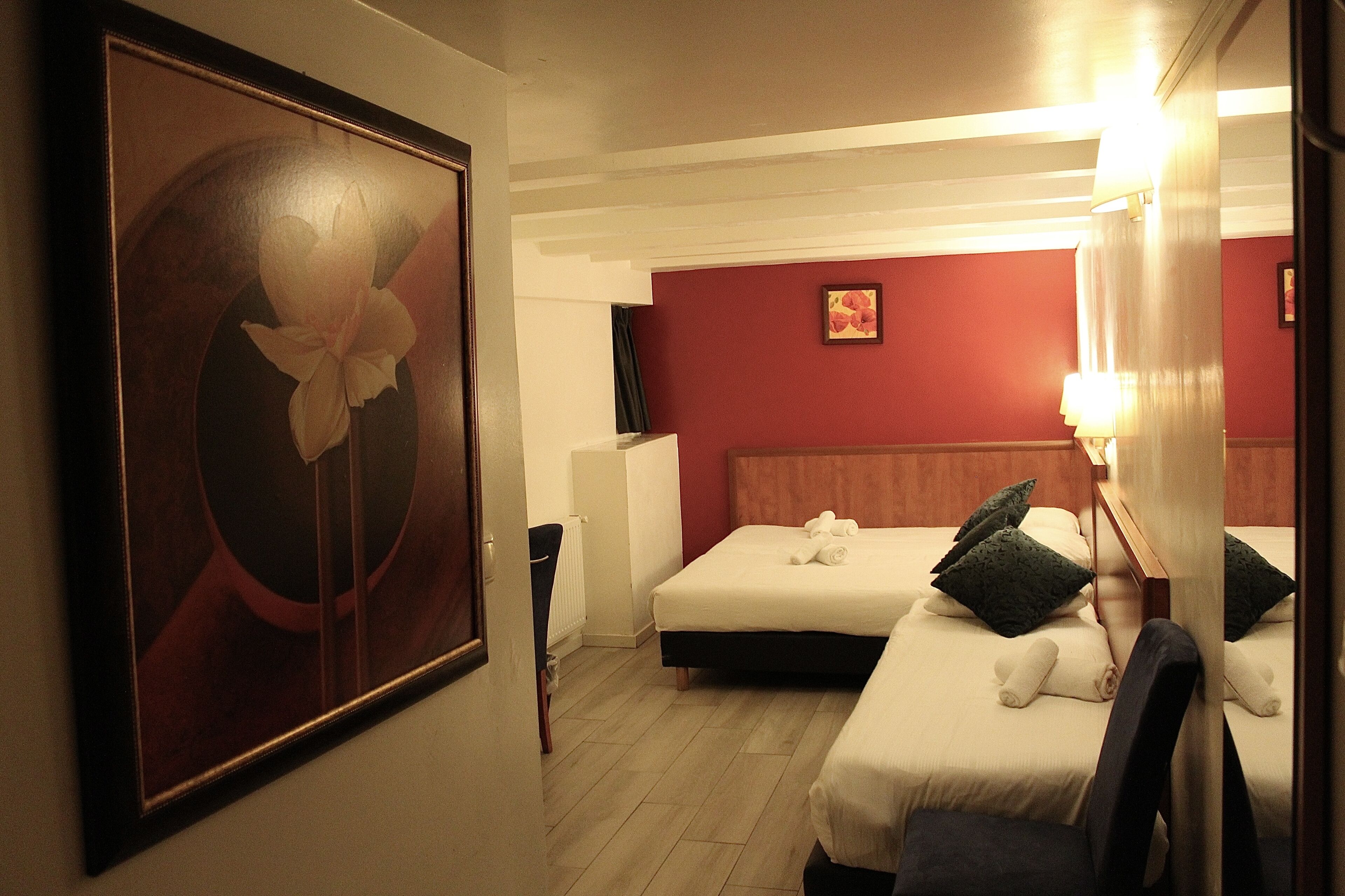 A hotel room with two beds, a large floral artwork on the wall, a red accent wall, and a bedside lamp.