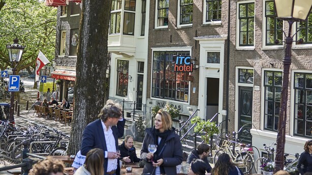 A bustling outdoor café with patrons seated at wooden tables, surrounded by a tree and a building with a visible 'ITC Hotel' sign. - ITC Hotel (Amsterdam)