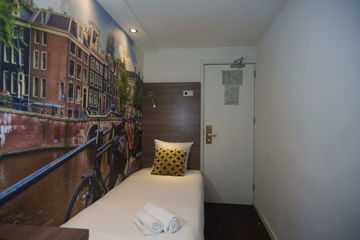 A hotel room with a bed, a wall mural of a cityscape, a door with a sign, and a towel on the bed.