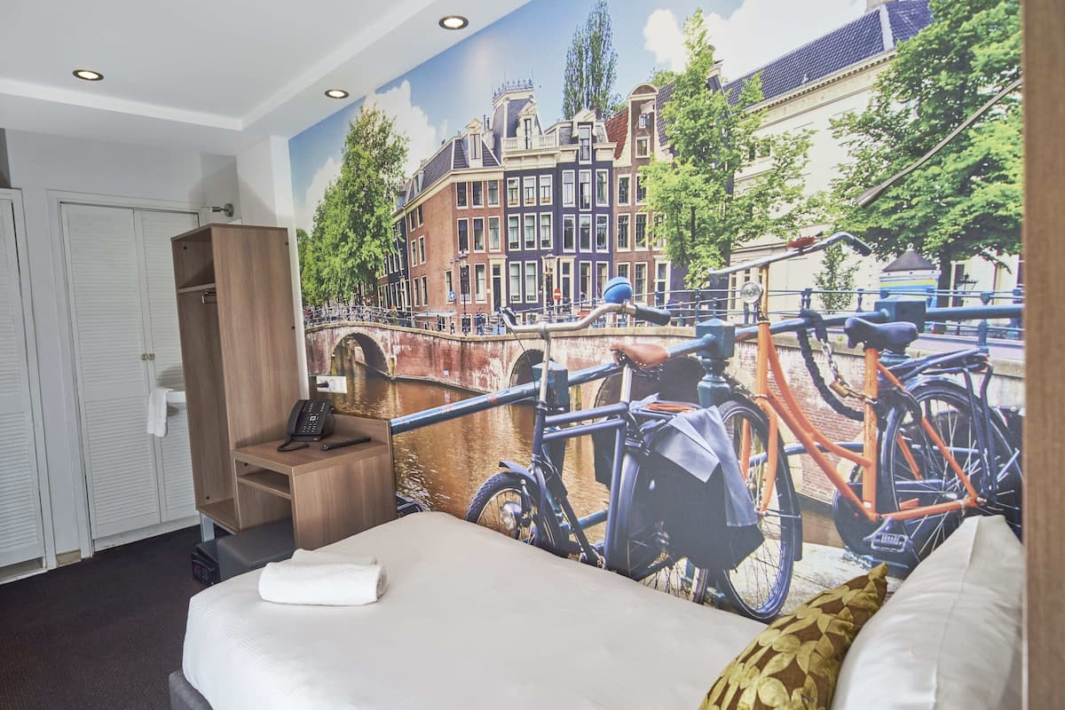 A hotel room with a mural of a canal scene, a bed, a wooden wardrobe, and a desk with a telephone.