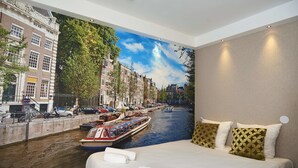 A hotel room with a mural of a canal, a bed with white linens, and two decorative pillows. - ITC Hotel (Amsterdam)