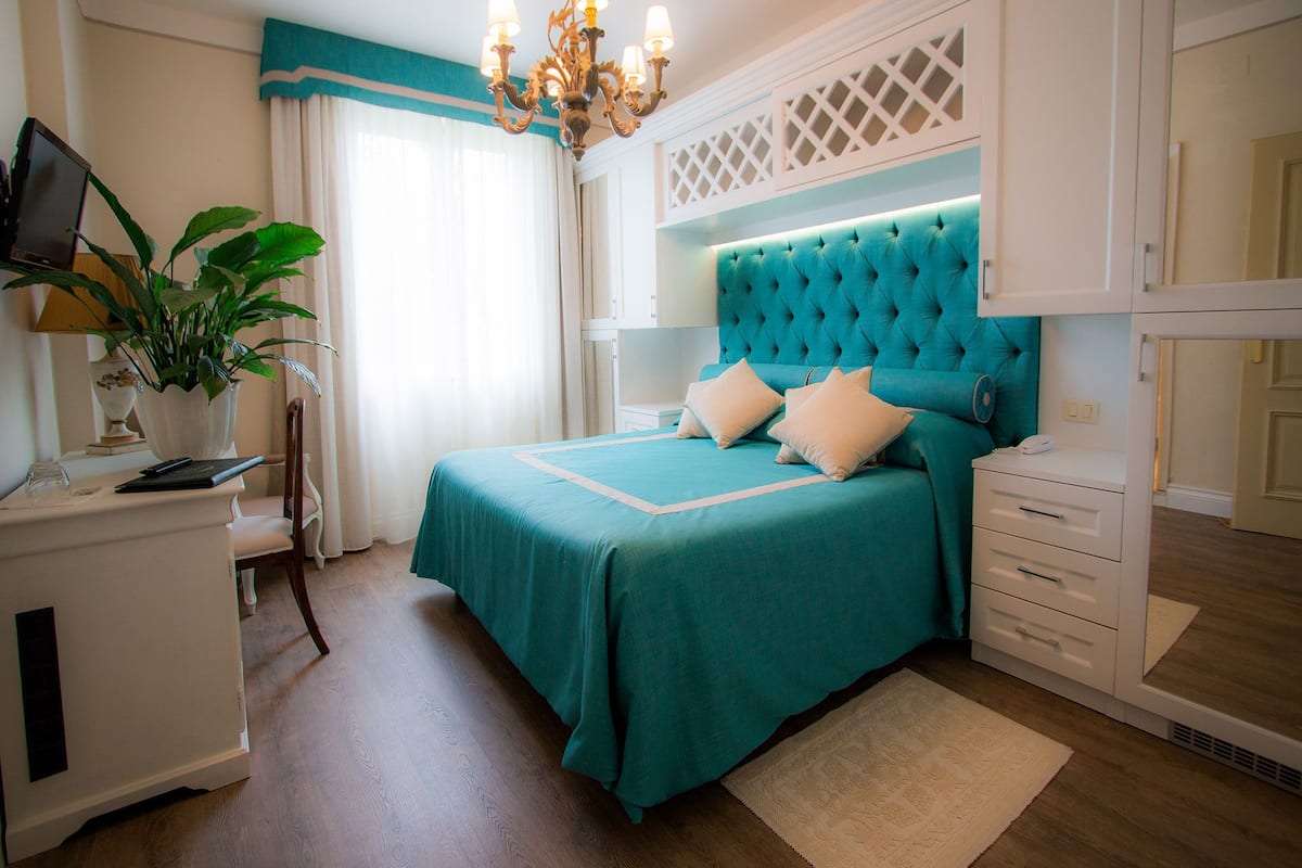 A bedroom with a teal bedspread, a white nightstand, a desk with a computer and a plant, a chandelier, and a large window with curtains.