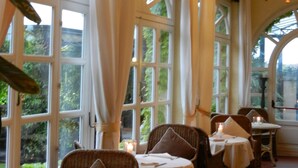 Un comedor con sillas y mesas de mimbre, ventanas amplias con cortinas y una araña.