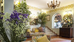 Un salón amplio con tapicería floral, un jarrón grande de flores y una escalera que conduce a un nivel superior.