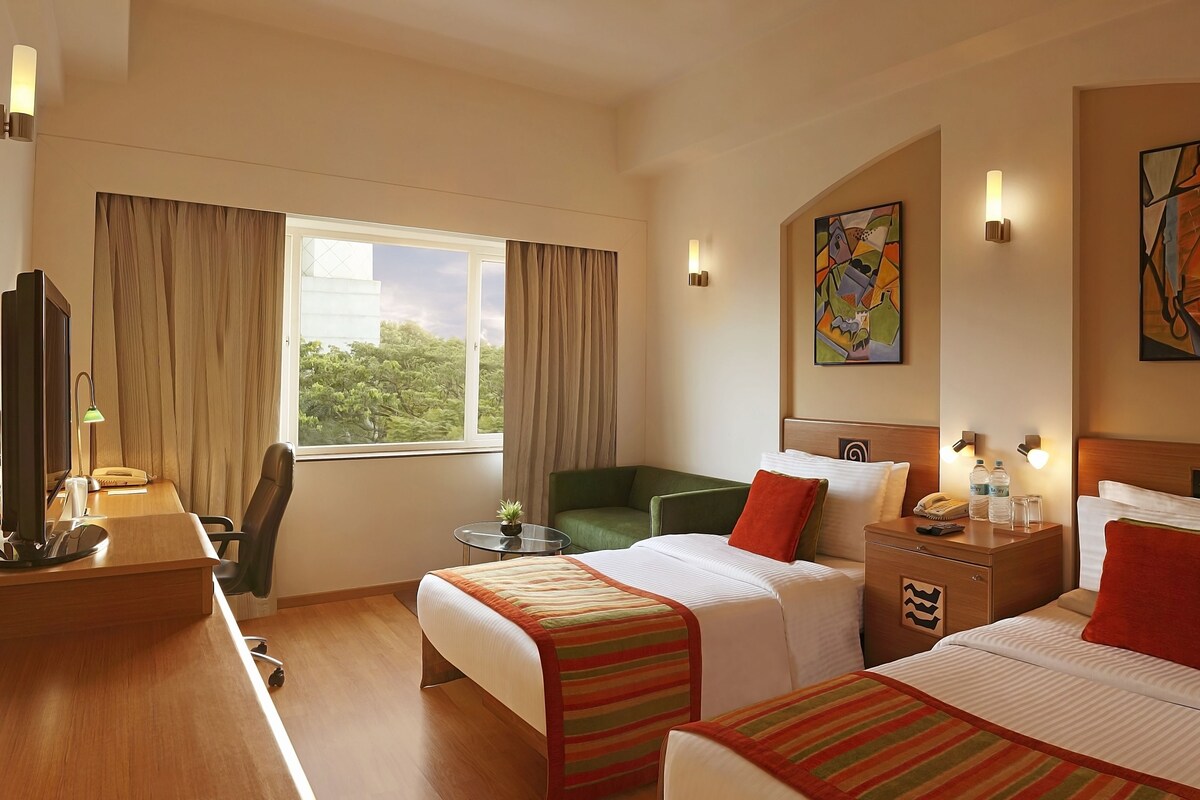 Habitación de hotel con dos camas, un escritorio, una silla, una mesita con una planta y obras de arte en las paredes.