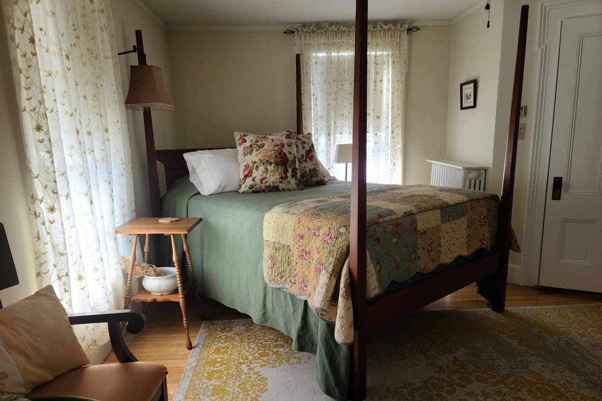 A four-poster bed with floral bedding, a wooden nightstand with a lamp, a green upholstered chair, and a patterned area rug.