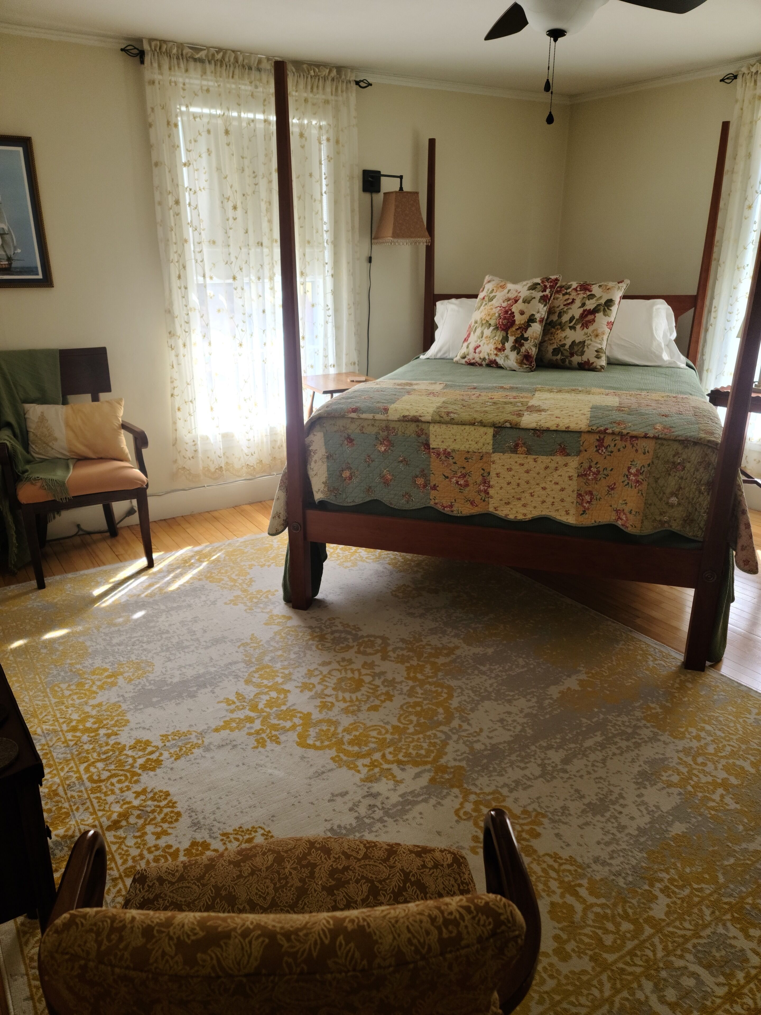 Una cama con dosel, ropa de cama floral, piso de madera y una silla con un cojín estampado.