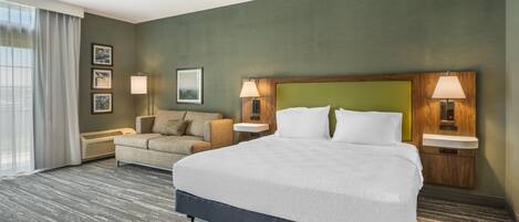 A hotel room with a large bed, a sofa, two bedside lamps, a wall-mounted TV, and a window with curtains.