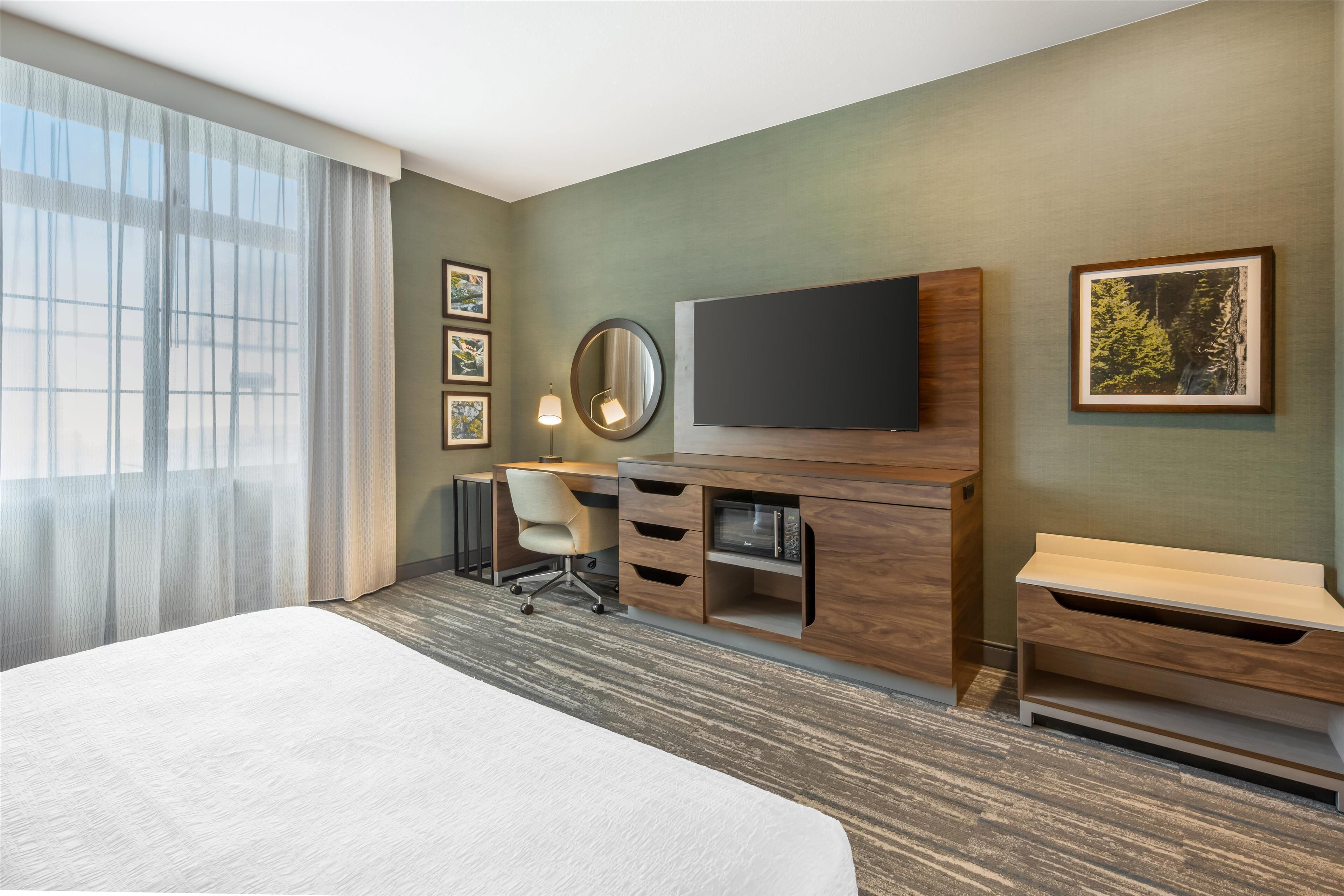 Une chambre d’hôtel dotée d’un grand lit, d’un bureau avec une chaise, d’un téléviseur à écran plat, d’un miroir et d’œuvres d’art accrochées aux murs.