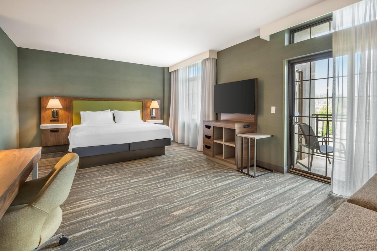 Habitación de hotel con una cama grande, un escritorio con silla, televisor y balcón con una silla.