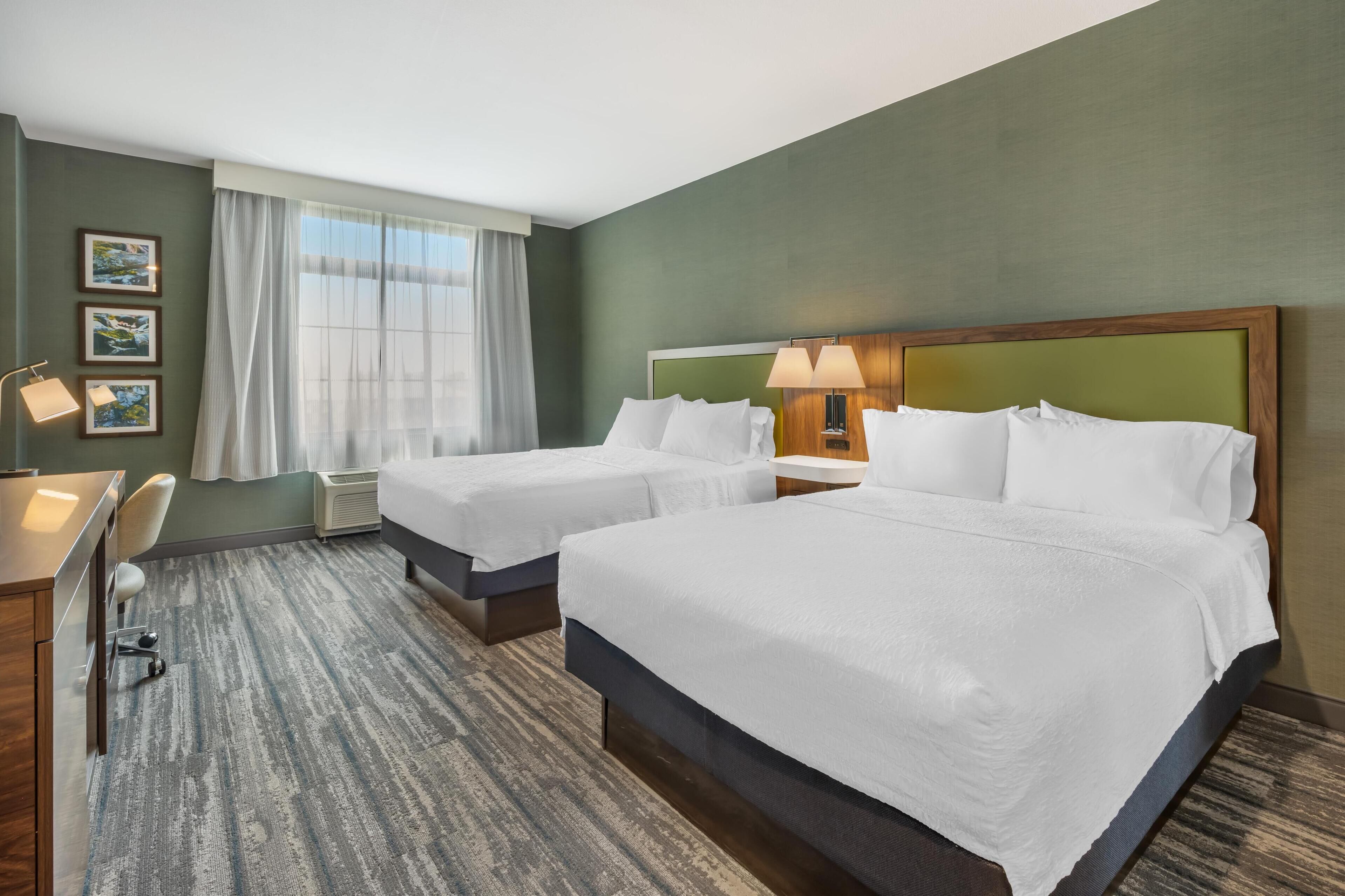 A hotel room with two beds, a desk, and a chair. There are framed pictures on the wall, a window with curtains, and a lamp on the desk.