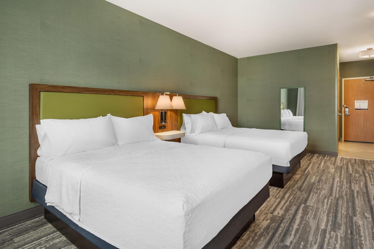 Una habitación de hotel con dos camas, una mesita de noche con lámpara, un espejo y una puerta.