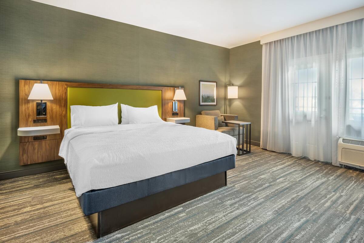 Habitación de hotel con una cama grande, dos mesitas de noche con lámparas, una pequeña mesa con una silla y un amplio ventanal con cortinas.