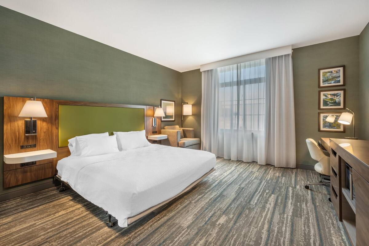 Una habitación de hotel con una cama grande, un escritorio con silla, una mesita pequeña y una ventana con cortinas.