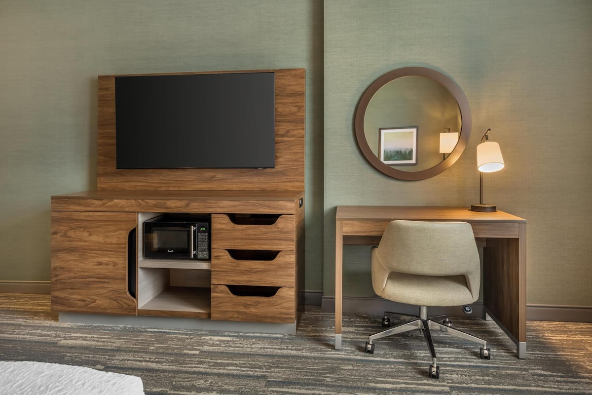 Una habitación de hotel con un escritorio de madera, un televisor de pantalla plana, una lámpara y un espejo.
