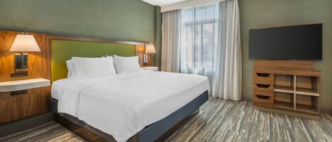 A hotel room with a large bed, two bedside lamps, a flat-screen TV, a wooden nightstand, and a large window with curtains.