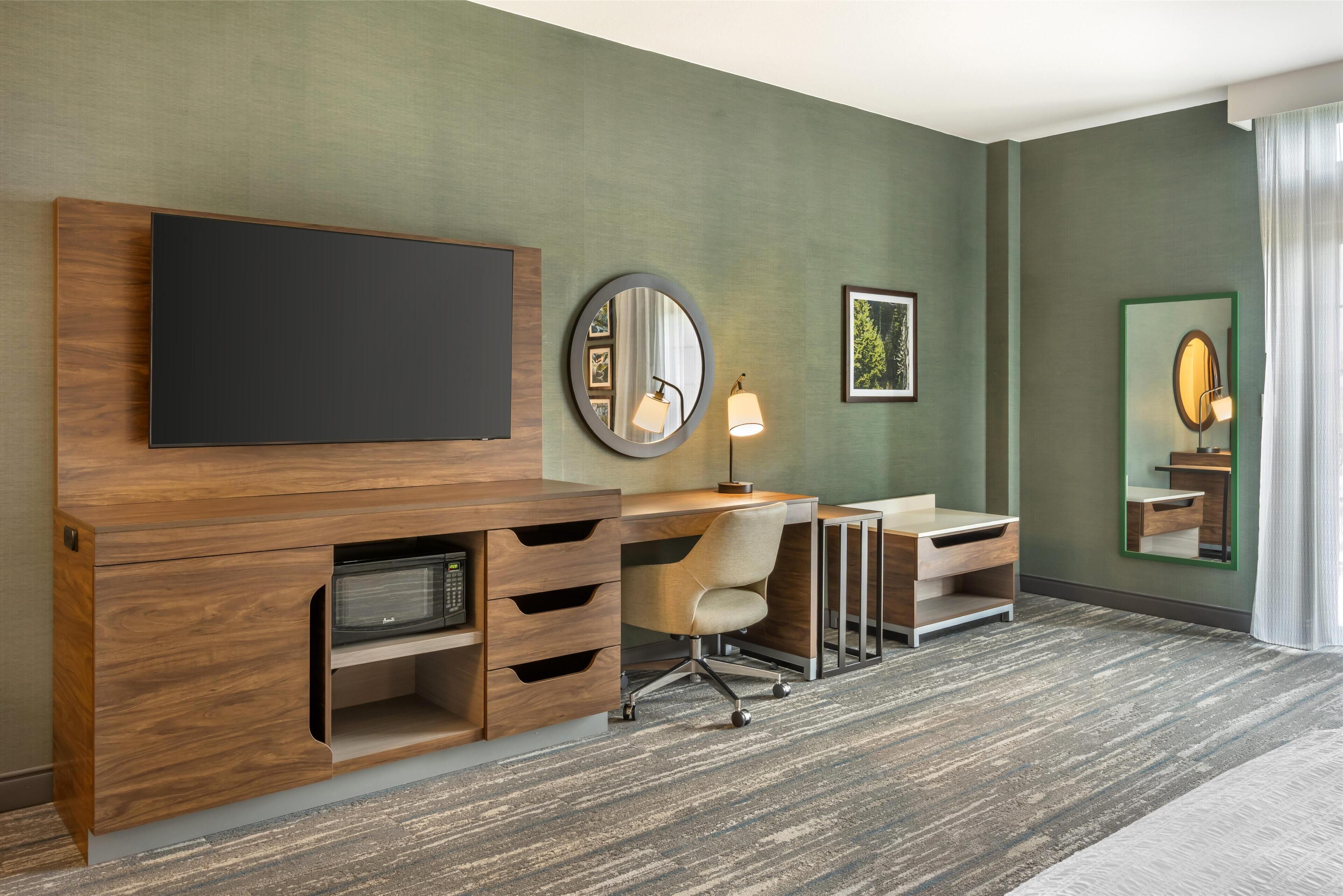 A modern hotel room with a flat-screen TV, a desk with a chair, a mirror, and a large window with curtains.