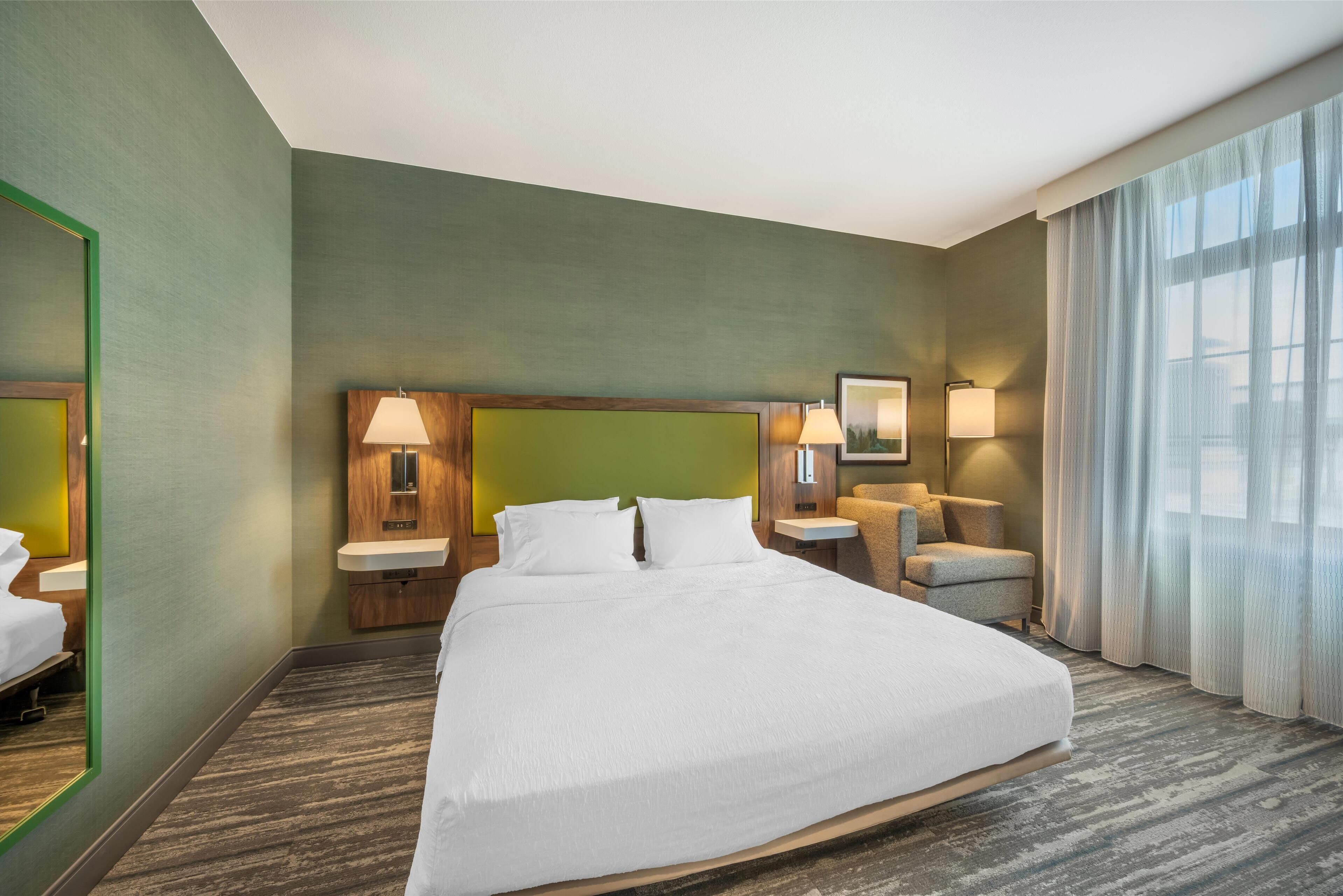 A hotel room with a large bed, a wooden headboard, a bedside table with a lamp, a chair, and a large window with curtains.