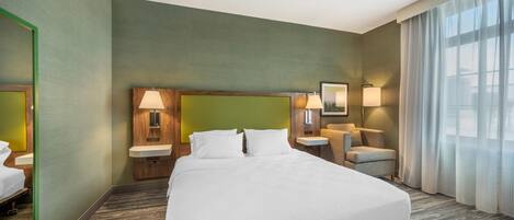 A hotel room with a large bed, a wooden headboard, a bedside table with a lamp, a chair, and a large window with curtains.