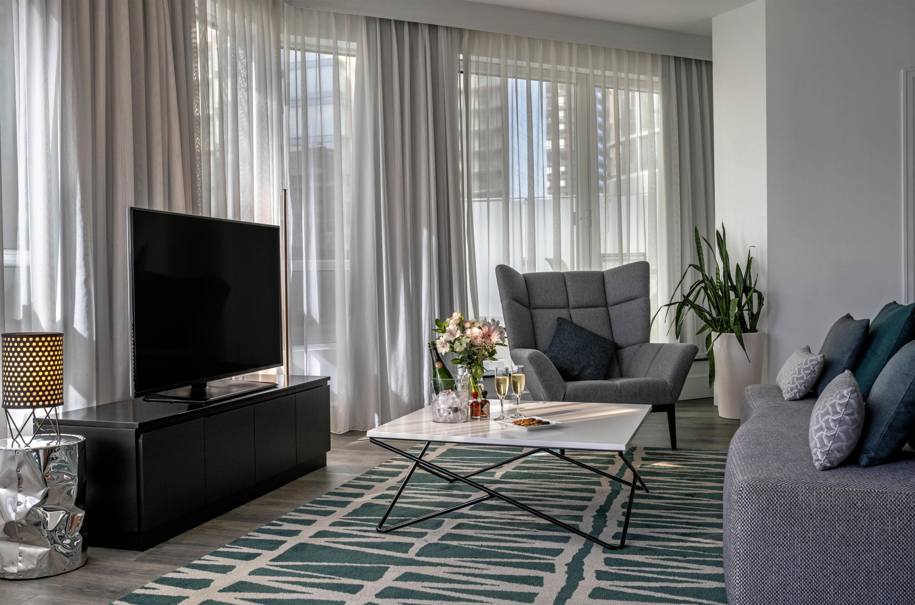 A modern living room with a flat-screen TV, a grey sofa, a coffee table with drinks and flowers, and sheer curtains.