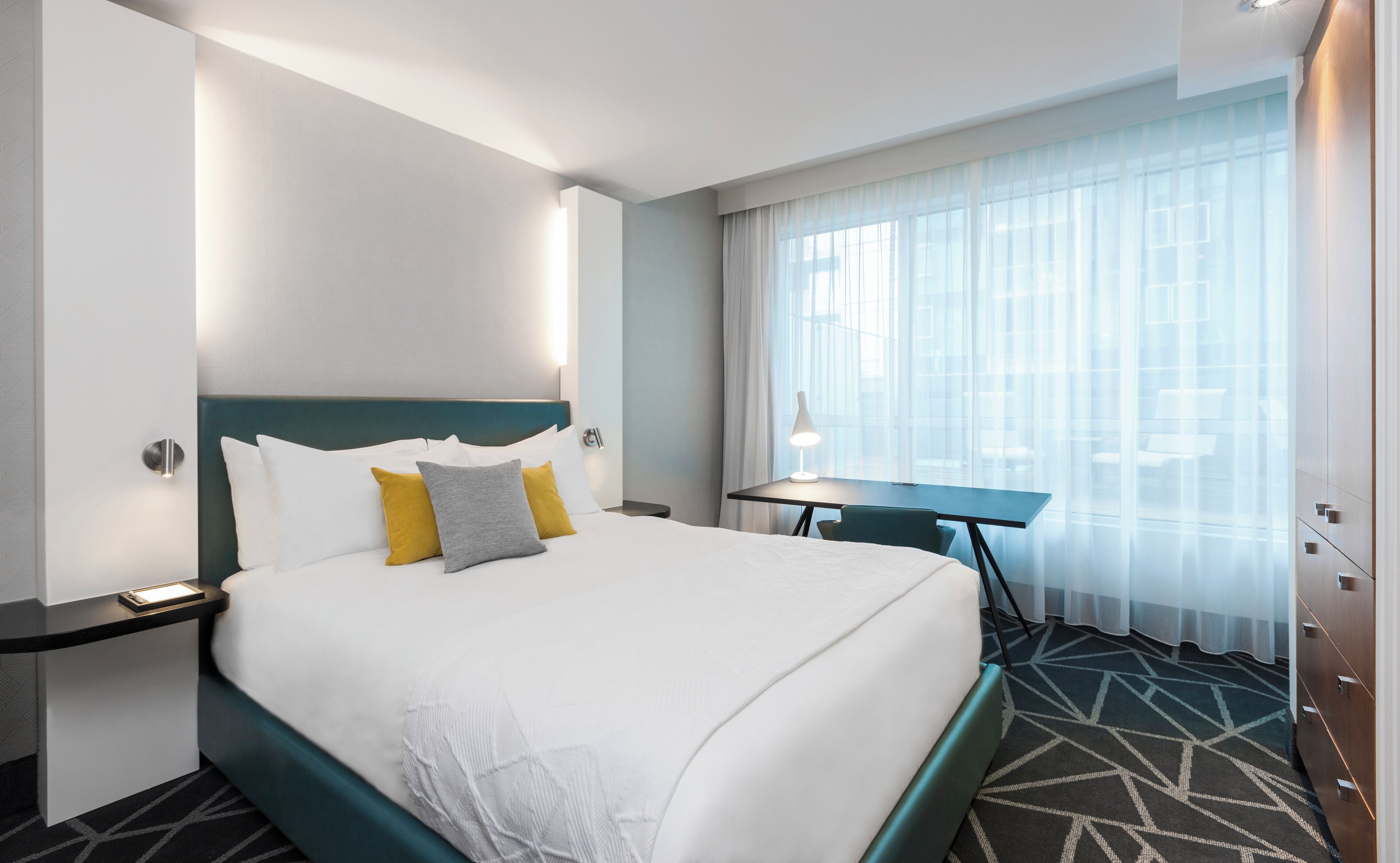 A hotel room with a large bed, a desk, and a view of the city through sheer curtains.