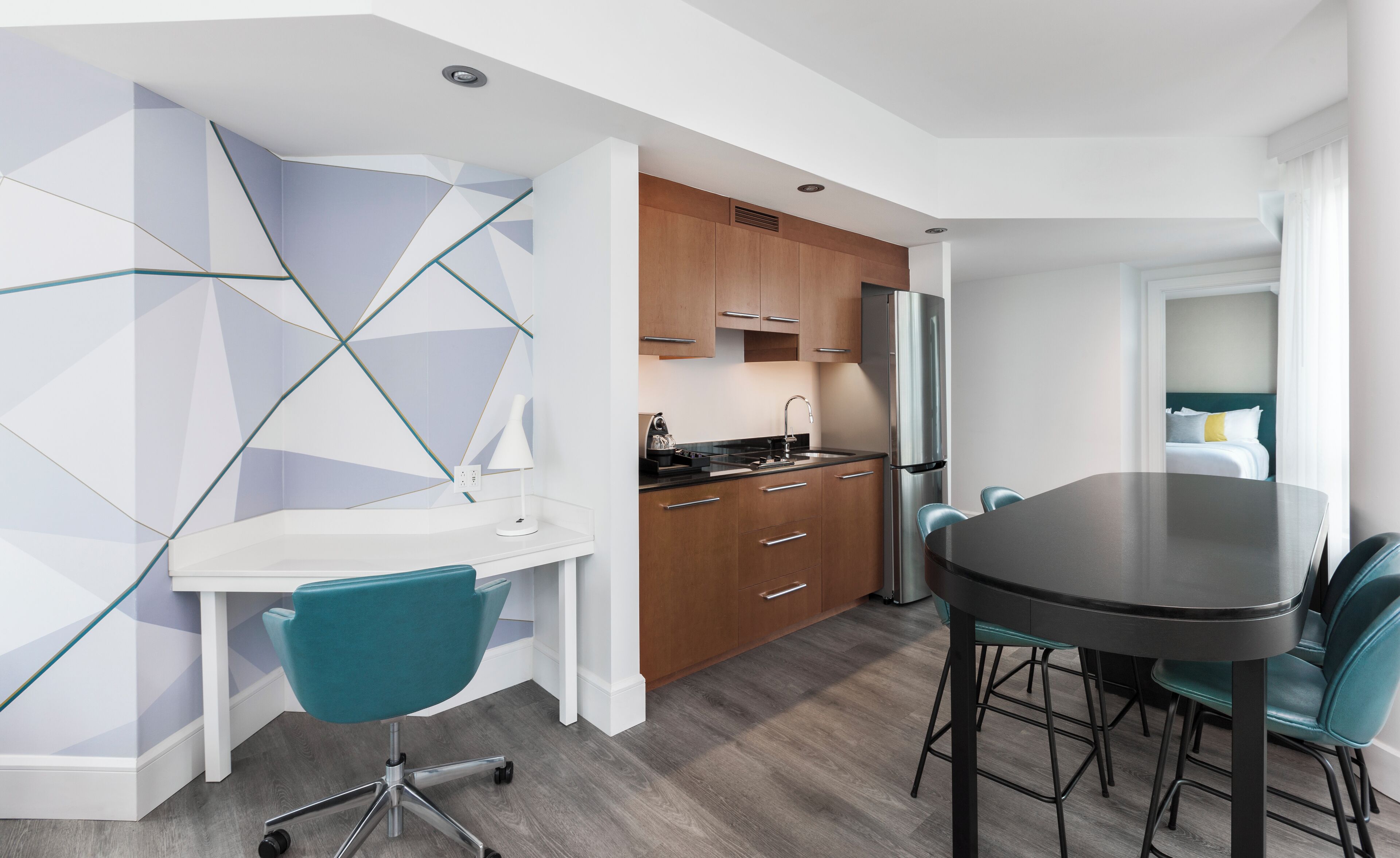 A modern kitchen with a white countertop, wooden cabinets, and a stainless steel refrigerator. There is a dining table with teal chairs and a white desk with a teal chair.