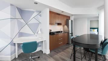 A modern kitchen with a white countertop, wooden cabinets, and a stainless steel refrigerator. There is a dining table with teal chairs and a white desk with a teal chair.