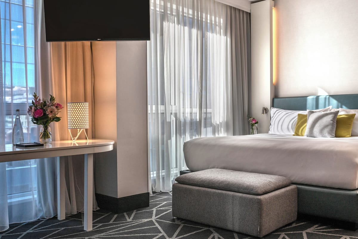 Habitación de hotel con cama, una mesita con un florero, una silla y un ventanal con cortinas.