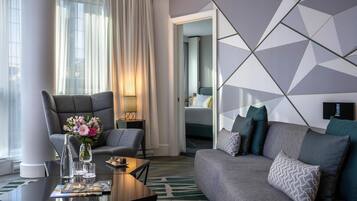 A modern hotel room with a grey sofa, a coffee table with a magazine, a vase with flowers, and a bed with a blue headboard.