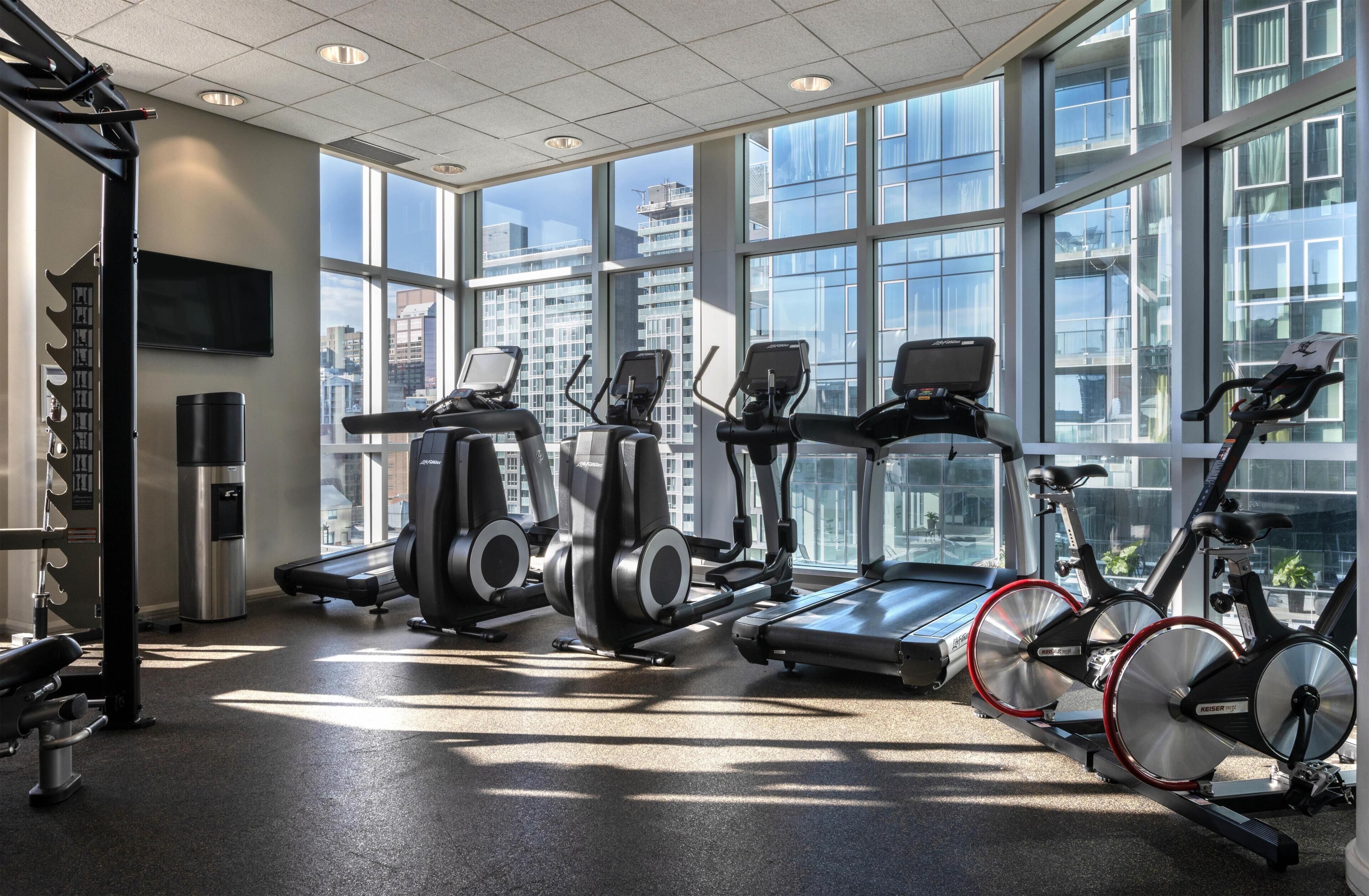 A gym with treadmills, elliptical machines, and a stationary bike, large windows, and a city view.
